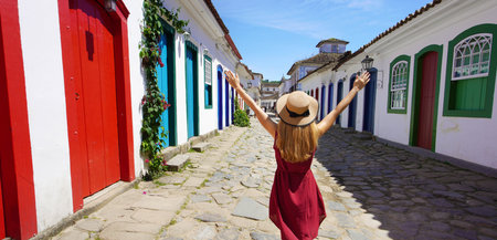 Joyful girl in Brazil. Beautiful young woman with raised arms walking in colorful historic town of Paraty, Brazil.の写真素材