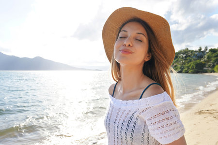 Happy woman with hat breathing and smiling at the beach on summer vacation. Beautiful young female with closed eyes enjoy nature relaxed outdoors. Happy lifestyle and well being concept.の写真素材