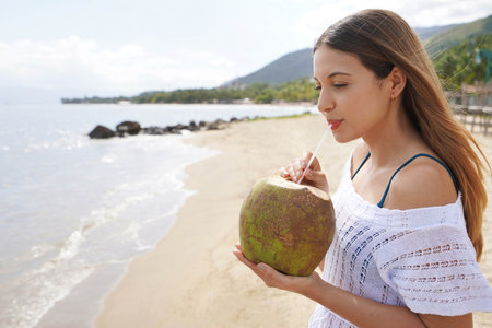 Portrait of young woman holding a fresh green coconut enjoying drinking on the beachの写真素材