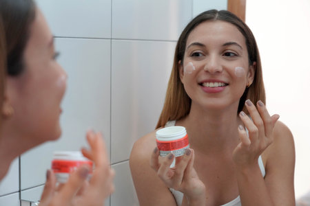 Close-up of relaxed young Brazilian woman applies moisturizer cream on her face after washing in bathroom. Enjoys beauty treatments. Uses cosmetic product for healthy skin.の写真素材