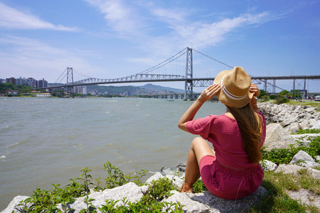 Young traveler woman in pinkish clothes and hat sitting on sea shore looking the Hercilio Luz...
