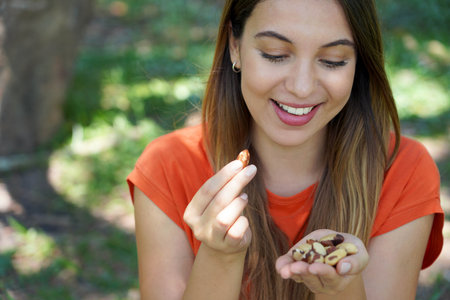 Close-up of healthy smiling woman eating Brazil nuts in the park. Looks at nuts.の写真素材