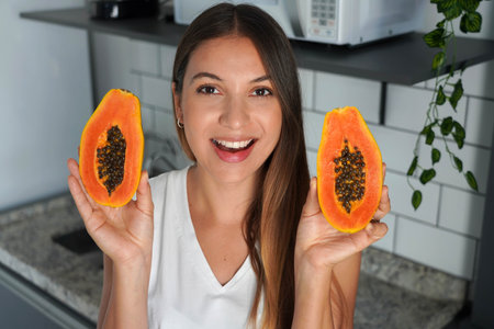 Beautiful young woman showing a papaya cut into two halves in the kitchenの写真素材