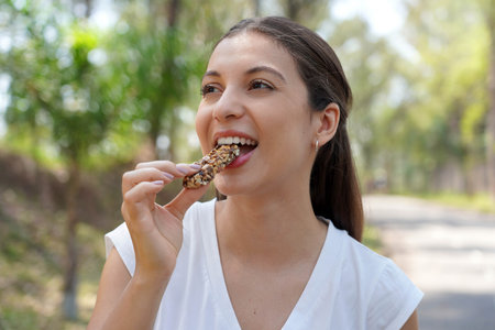 Beautiful young woman biting a nuts cereal energy bar in city parkの写真素材