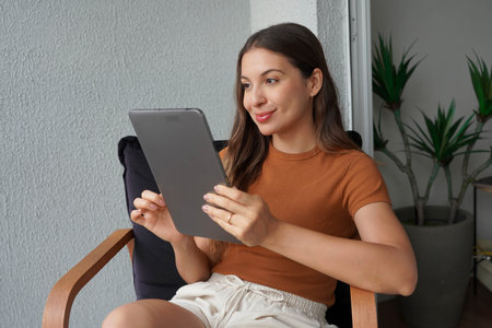 Multi-ethnic girl reading an e-book on tablet sitting relaxed on armchair at homeの写真素材