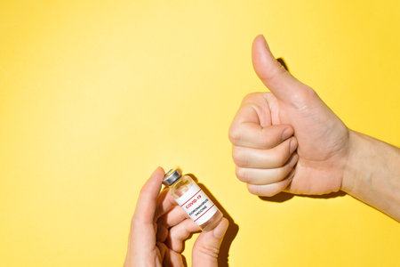 Male hands holding a bottle of covid-19 vaccine and showing a Like gesture on a yellow background. top view, copy spaceの写真素材