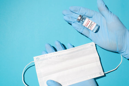 Doctor's hands in medical gloves, a bottle of covid-19 vaccine and a medical face mask, top view....