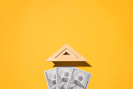 Wooden roof of toy house and one hundred dollar bills on yellow background, flat lay. Concept of buying a home, real estate cost, renting an apartment, investing in construction. top view, copy spaceの写真素材