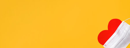 Red heart in a medical mask on a yellow background, flat lay. Conceptual banner, copy space.の写真素材