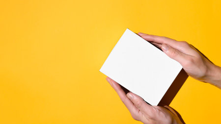 Male hands holding a box on a yellow background, flat lay. Empty white box mock up. Top view, copy space. Delivery concept, gift box.の写真素材