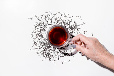 Female hand holding a cup of tea top view. Freshly brewed black tea and scattered dry ceylon tea leaves on a white plate.の写真素材