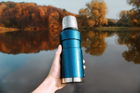 A man holds in his hand a blue flask, background of the autumn forest and lake. The concept of hiking, outdoor recreation, tourism.の写真素材