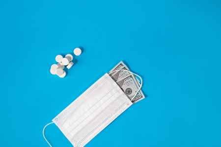 Pills and medical face mask covered one hundred dollar bill on blue background. Concept of economic crisis caused by coronavirus pandemic, expensive treatment and vaccine, the pharmaceutical business.の写真素材