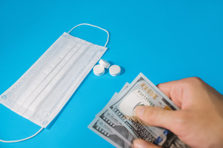 Person holds money in hand near a medical mask and medication. It symbolizes the purchase of medicines, the cost of treatment, medicine and healthcare, the price of a vaccine.の写真素材