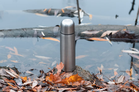 Vacuum steel flask close-up, outdoors. Hiking flask stands by the water. Hike concept, relaxation in nature, hot drink in cold weather.の写真素材