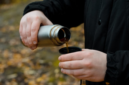 Man pours a hot drink from a flask into a mug outdoors. Close-up male hands holding a hiking flask against the background of an autumn forestの写真素材