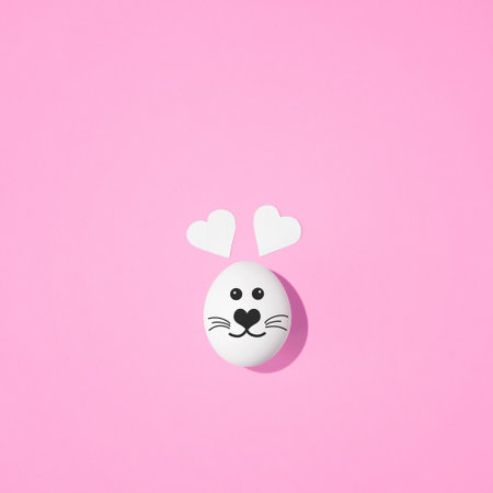Creative minimal Easter concept. Rabbit face painted on egg with heart-shaped ears on pastel pink background. flat lay.の写真素材