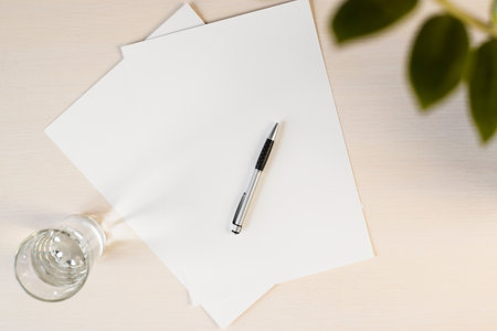 Blank paper, glass of water and pen on the table. Flat lay mock up. Home plant in the frame. copy...