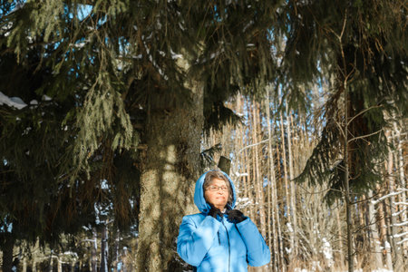 Senior woman standing in pine forest winter. Smiling pensioner enjoying sunny day. Walk in the winter forestの写真素材