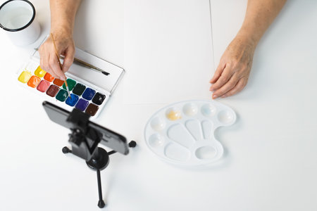 An elderly woman's hands paint with watercolors indoors, close-up. Smartphone, paints, brushes on the table top view. Online drawing course, retirement hobby conceptの写真素材