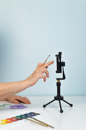 Woman's hand holding a paintbrush and using a phone on a tripod. Blog, online broadcast to social...