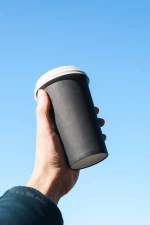 Male hand holding disposable cup of coffee against blue clear sky, close-up. Vertical photo,...