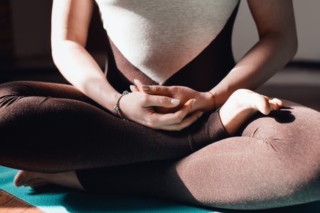 Young woman in tracksuit practicing meditation sitting in lotus position on yoga mat, indoors at sunrise. Close-up. Concept of mindfulness meditation, healthy lifestyleの写真素材