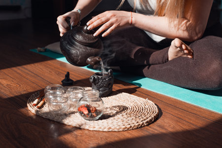 Woman sitting in lotus position, making herbal tea, close-up. Tea ceremony in the yoga and meditation room at sunrise. Clay teapot, wooden spoons on a tray and rising steamの写真素材