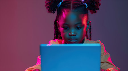 Small child sits in front of a laptop and engages in online learning and educational activities. An African American girl looks focused and attentive while interacting with digital lessons on screen.の素材