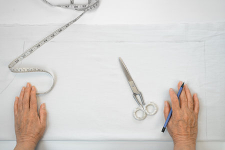 Hands of an elderly woman designer clothes, top view. Women seamstresses, pencil, white fabric,...