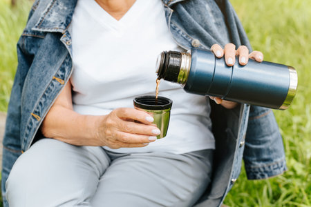 Outdoor camping. Senior woman having a rest in nature. Elderly woman's hands holding a flask and pouring tea into a mug. Cropped imageの写真素材