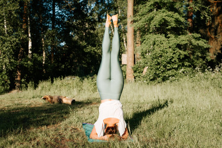 Slender young woman practicing yoga asana headstand back view, outdoors. Sportive girl exercising on a yoga mat in nature in the morning, outside. Healthy active lifestyle concept.の写真素材