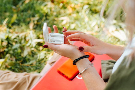 Close-up of young woman and wireless earphones outdoors. Selective focus on female hands holding box and taking white bluetooth headphones. Hipster teenage girl using modern audio device.の写真素材