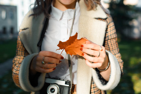 Close-up young woman hipster photographer with retro film camera holding orange fallen leaf outdoors. Selective focus on female hands and maple leaf. Hello October autumn lifestyle.の写真素材