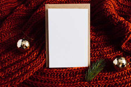 Christmas greeting vertical card mock-up for festive invitation or xmas wish. Happy new year or merry christmas composition layout. Golden balls, pine twig, cozy red knitted background, craft envelopeの写真素材