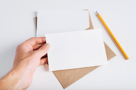 Postcard layout. Close-up of hand holding blank white card with empty space for branding. Craft envelopes, pencil on white table.の写真素材