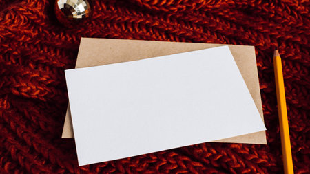 Merry christmas or happy new year composition mock-up, greetings card with blank space for festival invitation, xmas wishes. Christmas ball, pencil, craft envelope, white card on knitted red backgroundの写真素材