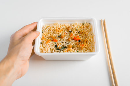 Close-up of hand holding box with ready-made instant Chinese noodles. Popular Asian dish and chopsticks on white table indoors.の写真素材