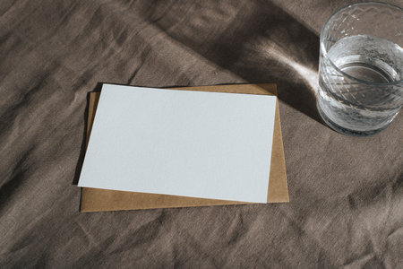 White card mock up with empty space for message, craft envelope and glass of water on rustic textile background with shadows. Close-up postcard layout.の写真素材