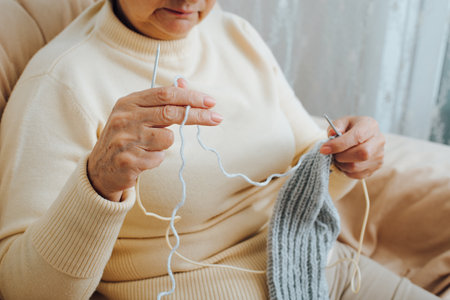 Close-up wrinkled hands of senior woman practicing needlework, handcraft, retirement active leisure. Grandmother knitting scarf while sitting on sofa indoors. Selective focus on front hand.の写真素材