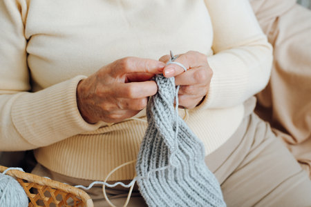 Close-up of senior woman's wrinkled hands knitting scarf, holding needles and threads. Hobby...