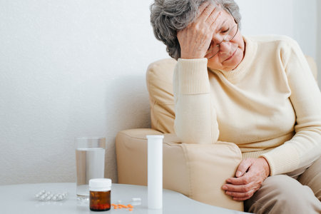 Senior woman with glasses with headache sitting in chair by table with bottles of medicines and pills, indoors. Elderly and healthcare, disease, pain, treatment concept.の写真素材