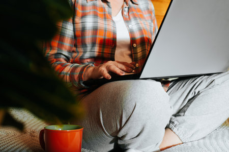 Close-up woman typing on laptop with tea or coffee mug while sitting on sofa at home. Freelance, coaching, remote office, distance work, e-learning concept. Selective focus on female hands.の写真素材