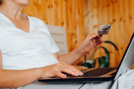 Side view of woman typing on laptop and holding bank credit or debit card while sitting on bed indoors, close-up. Online shopping, e-commerce concept. Selective focus in the foreground.の写真素材