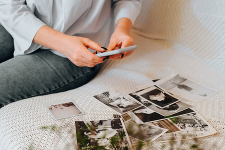 Close-up of caucasian woman photographing old black-and-white photos from family album on smartphone camera while sitting on sofa, indoors. Selective focus on female hands holding mobile phone.の写真素材