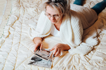 Nostalgia and memories concept. Middle-aged caucasian woman looking at old family album photos while relaxing on bed in bedroom, top view. Adult woman and vintage pictures of childhood and youth.の写真素材