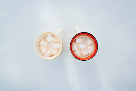 Two mugs of hot chocolate cocoa with marshmallows standing in snow, outdoors. Top view warming...