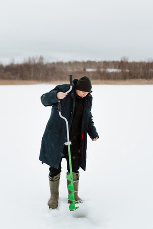 Winter fishing, fisherman drilling hole in ice while standing in snow on frozen water, outdoors. Winter hobby, active leisure, sport concept.の写真素材