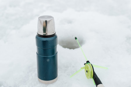 Winter fishing, flask and small fishing rod near drilled hole in frozen water, close-up. Selective...