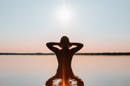 Silhouette of slim young woman swimming in lake at sunset, standing on water and enjoying outdoor recreation in nature, back viewの写真素材